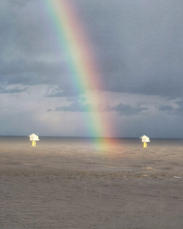 Bright rainbow descending into the sea under dark storm clouds, with two offshore structures visible on the water.
