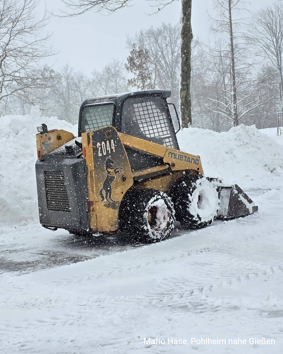 Ein Radlader räumt bei starkem Schneefall eine verschneite Fläche frei. Hohe Schneemengen liegen am Rand.