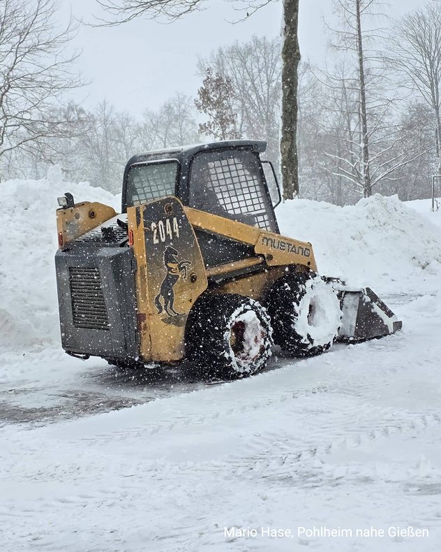 Ein Radlader räumt bei starkem Schneefall eine verschneite Fläche frei. Hohe Schneemengen liegen am Rand.