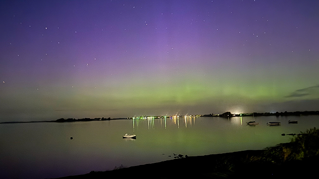 Grün-violette Polarlichter über Fehmarn, mit Spiegelungen im Wasser und Booten am stillen Ufer.