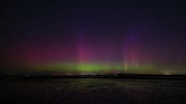 Polarlichter in grün, violett und rot über Husum, mit klar sichtbarem Sternenhimmel und dunklem Watt im Vordergrund.