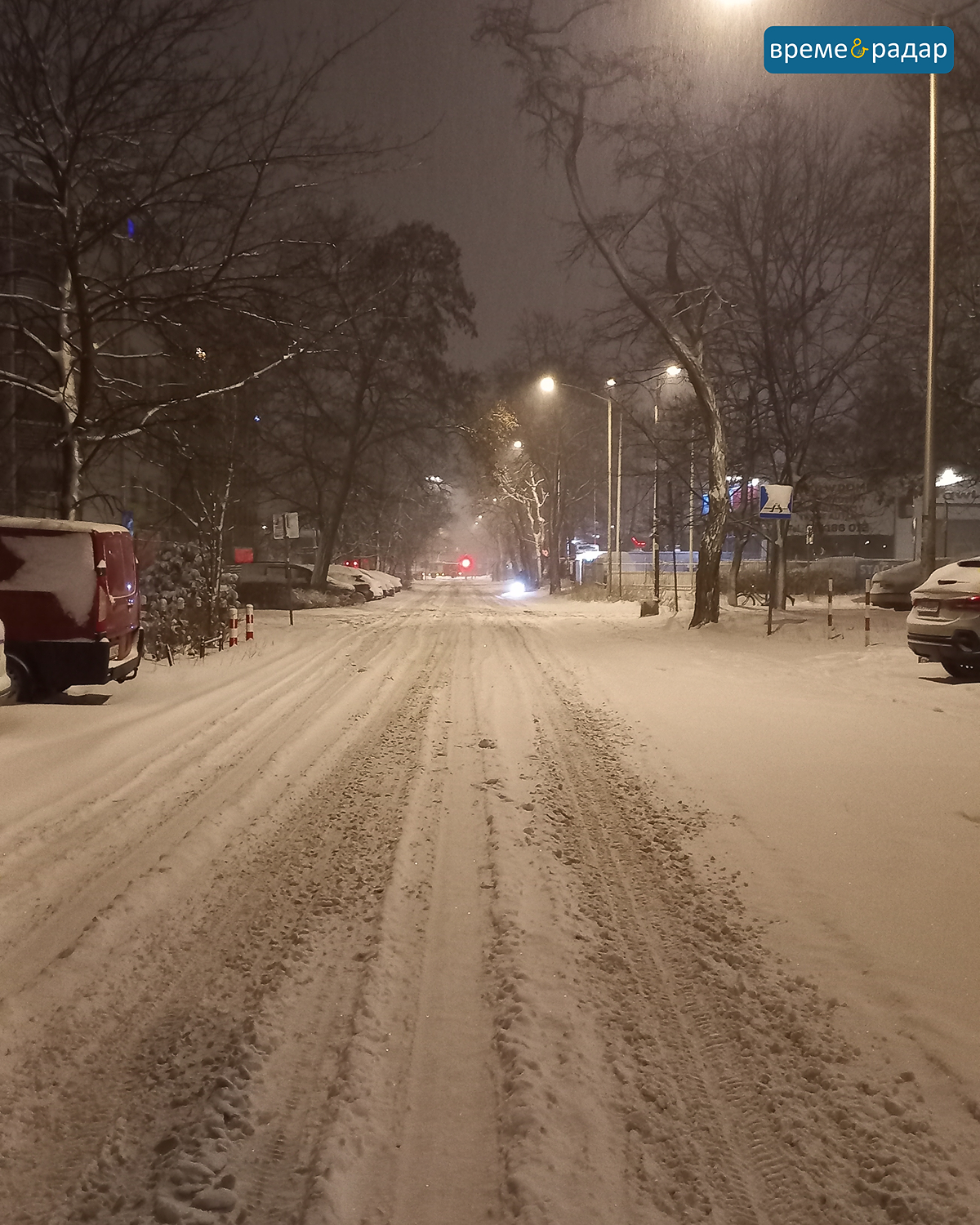 Заснежена улица с автомобилни следи, осветена от улични лампи през нощта.