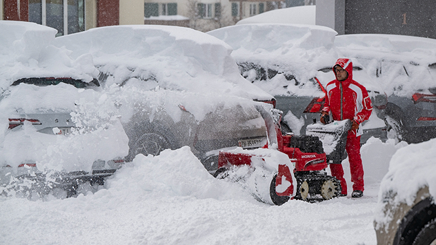 The picture shows a person with a snow blower. There is a lot of snow on the roofs of cars.