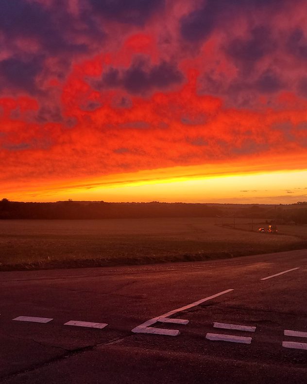 Intense red and orange sunset clouds glow above open fields and a rural road, with a bright yellow band along the distant horizon.