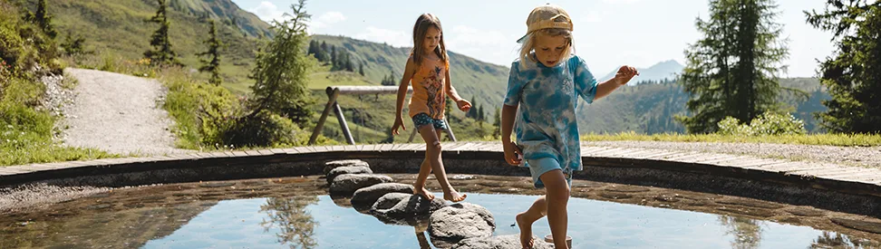 Zwei Kinder balancieren auf Steinen im Wasser.