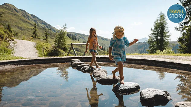 Zwei Kinder balancieren auf Steinen im Wasser.