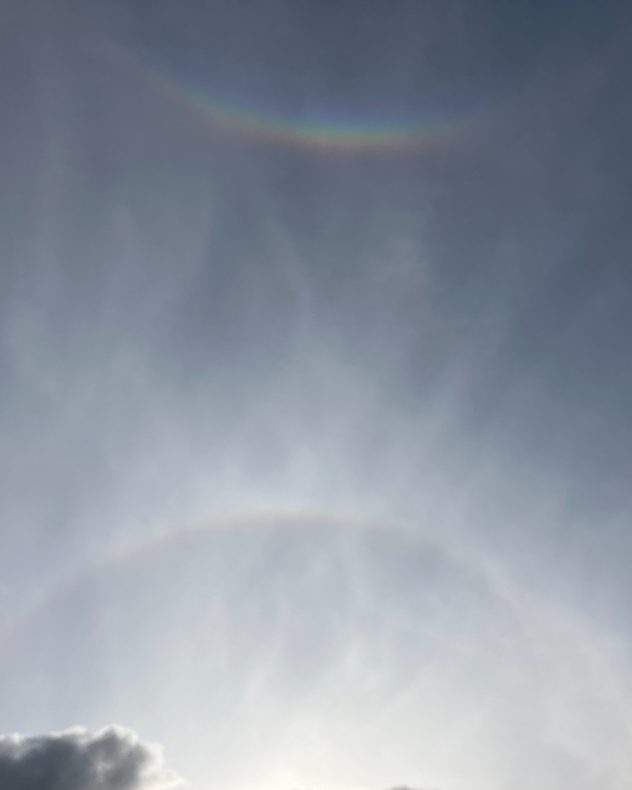 Faint rainbow-coloured circumzenithal arc high in a hazy gray sky, with a pale circular halo visible around the bright Sun below.