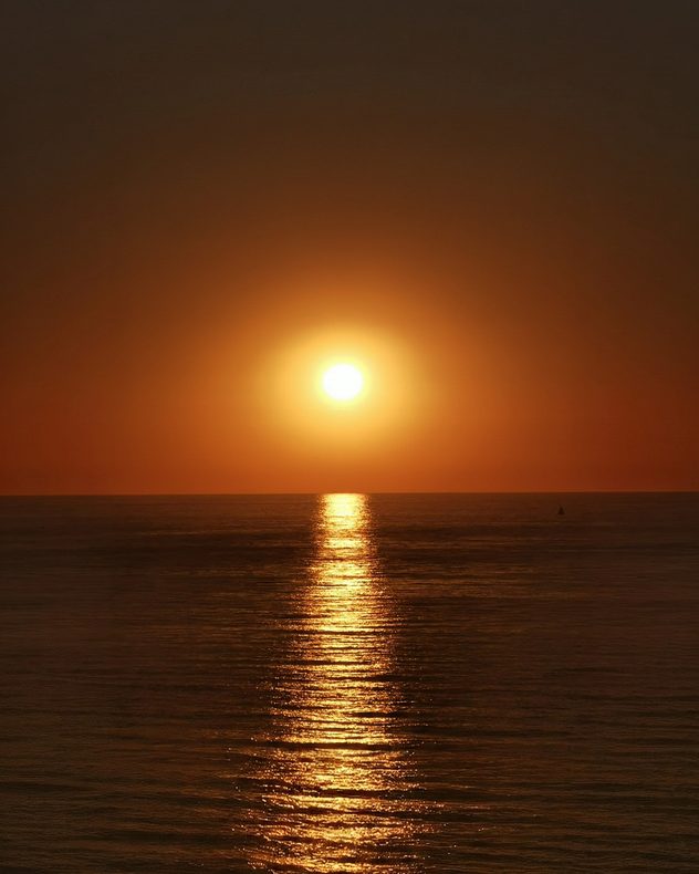 Bright golden Sun above a calm sea at sunset, casting a long shimmering reflection across the water beneath a clear orange sky.