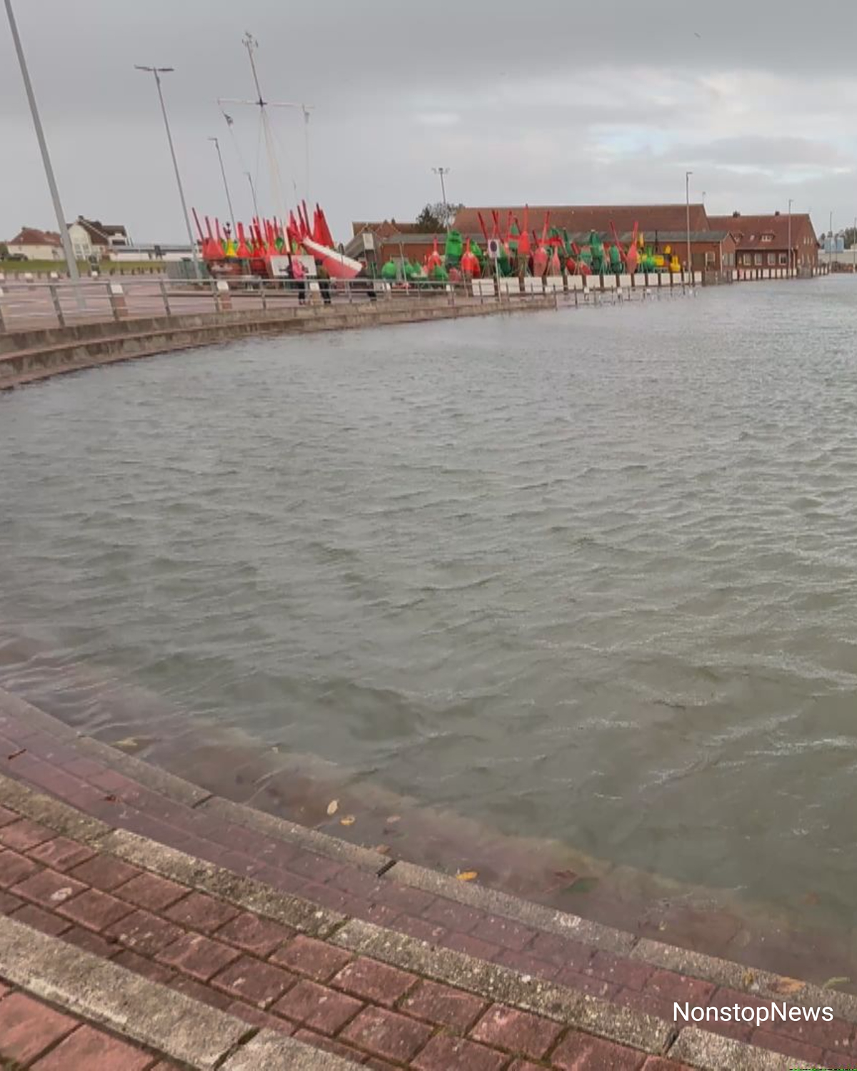 Überflutete Kaimauer an einem Hafen mit hohem Wasserstand, Bojen und Gebäude im Hintergrund unter grauem Himmel.