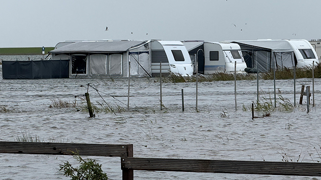 Überfluteter Campingplatz mit Wohnwagen und Vorzelten, Wasser steht hoch, grauer Himmel und Sturm an der Nordsee.