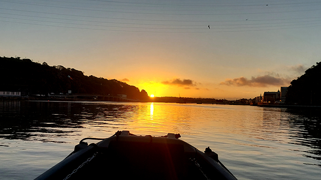 View from a boat on a calm river at sunrise, golden light reflecting on the water between two dark hills.