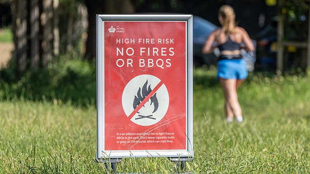 Red warning sign in a grassy park reading “HIGH FIRE RISK – NO FIRES OR BBQS,” with a crossed-out flame symbol, while a person walks in the background on a sunny day.