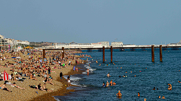 Busy pebble beach packed with sunbathers and swimmers along a crowded coastline, with people enjoying warm weather beside the sea and a long pier visible in the background.