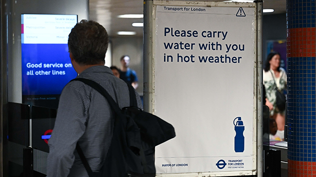 Commuter walking through a London Underground station beside a Transport for London sign reading “Please carry water with you in hot weather,” promoting hydration during high temperatures.