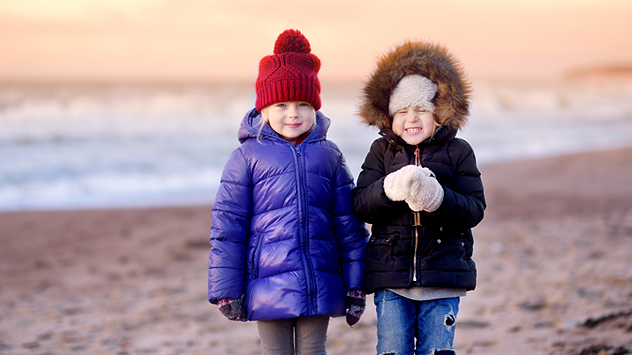 Zwei Kinder stehen warm eingepackt mit Winterjacken und Mützen am Strand, im Hintergrund das Meer bei Sonnenuntergang – es ist frühwinterlich kalt.