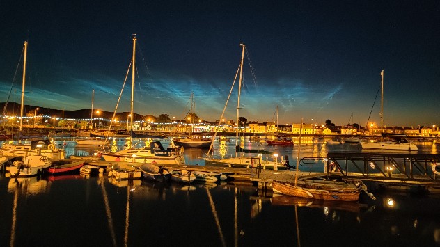 Nighttime marina with sailboats and glowing harbor lights beneath electric-blue noctilucent clouds stretching across the dark twilight sky.