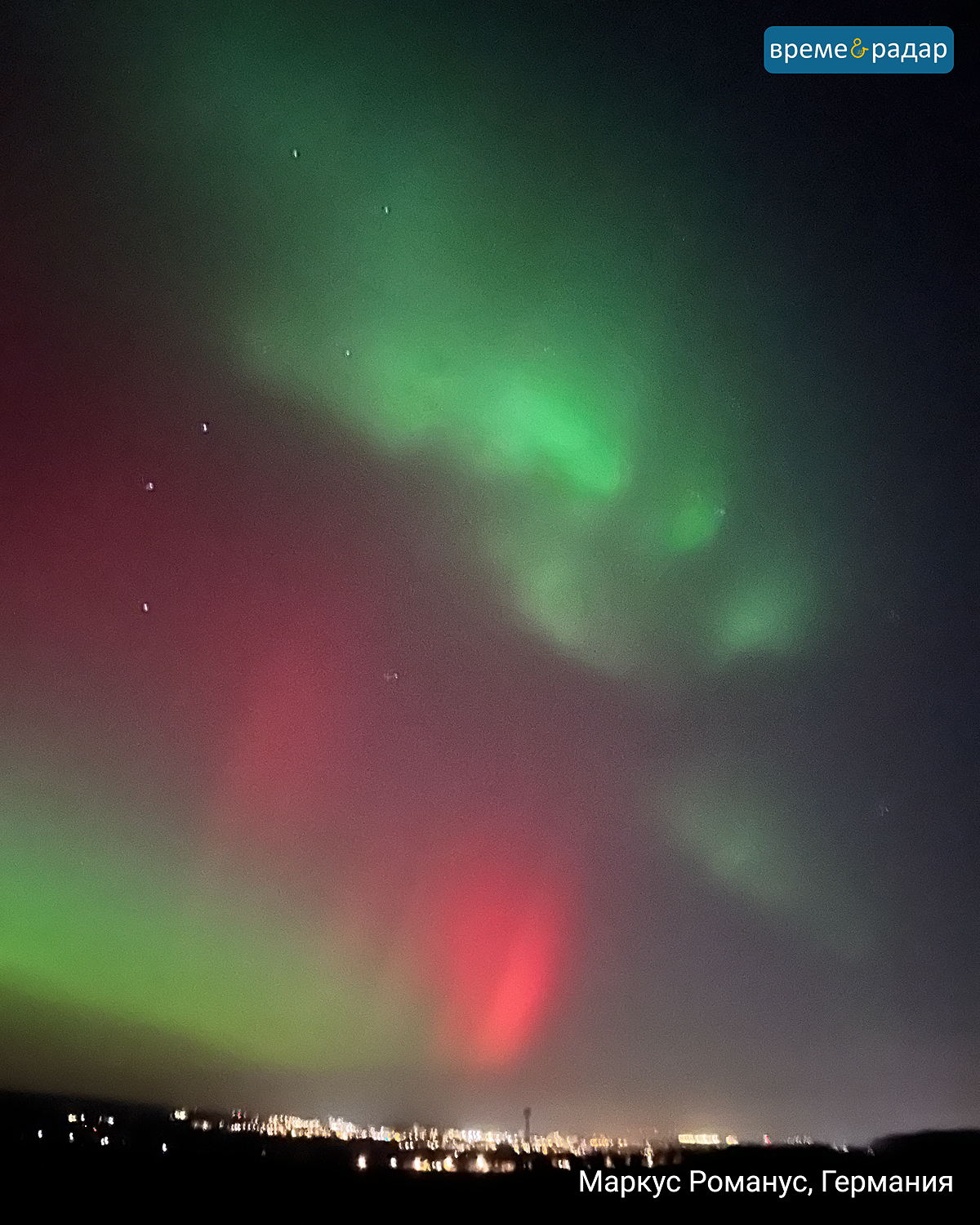 Зелено-червени полярни светлини над германски град през нощта. Светлините на града се виждат на хоризонта.