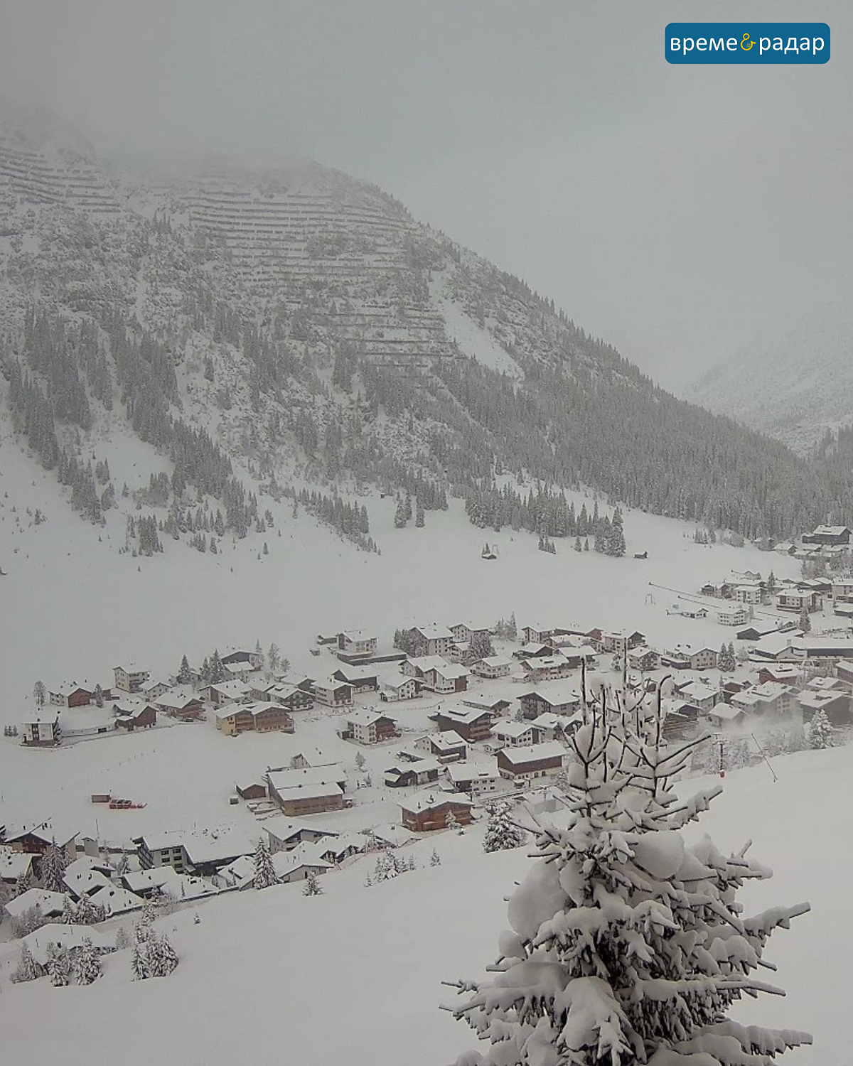 Заснежено алпийско селище с облачни планини и заснежени ели на преден план.