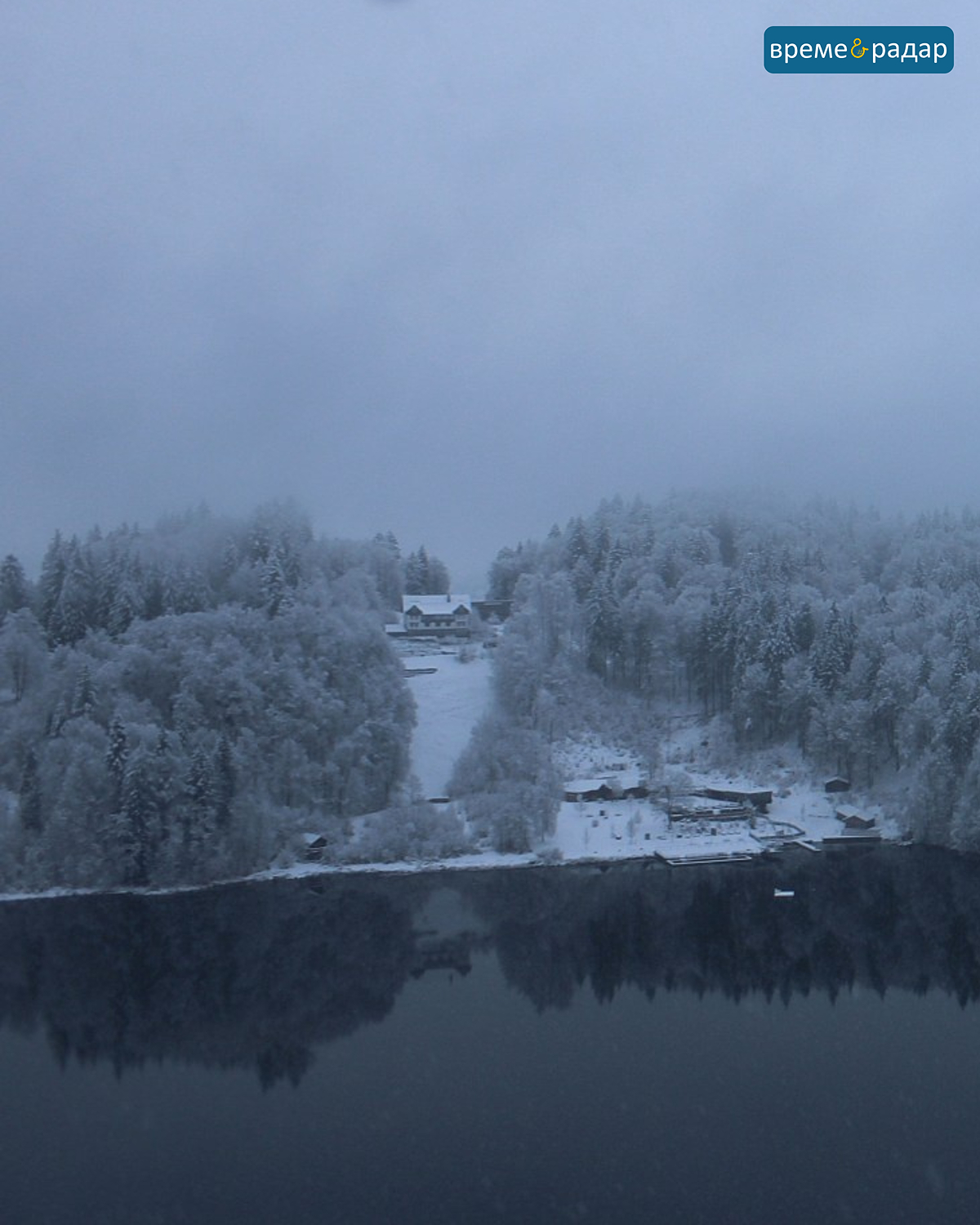 Заснежен склон с къща на тихо езеро, заобиколено от замръзнали гори. Лека мъгла покрива зимния пейзаж.