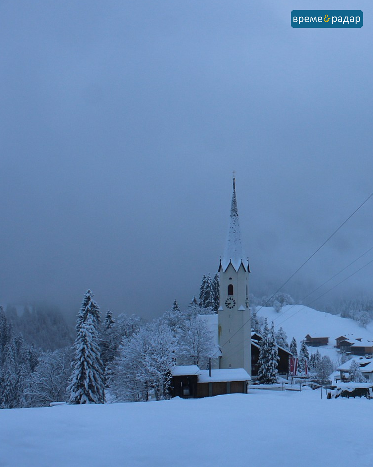 Заснежена църква в мъглива зимен пейзаж с къщи и дървета. Спокойна сцена в гъстата прясна снежна покривка.