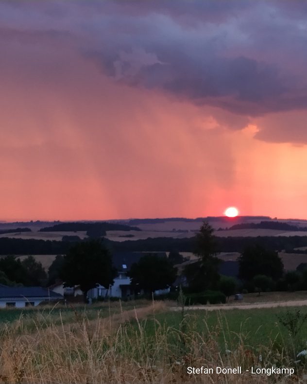 Die tief stehende Sonne leuchtet am Horizont, während ein Regenschauer aus dunklen Gewitterwolken über eine hügelige Landschaft niedergeht.