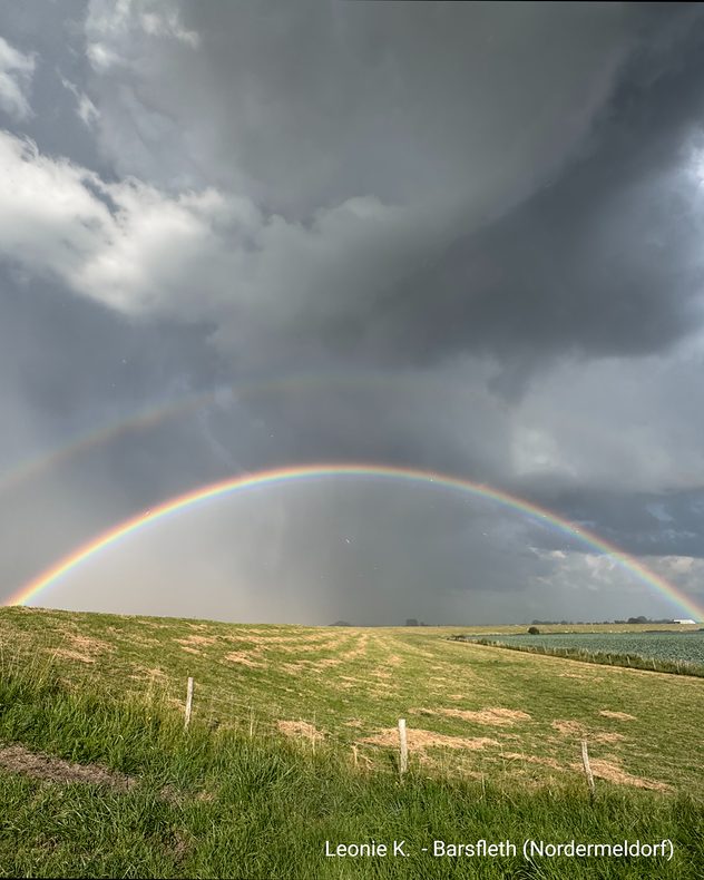 Ein kräftiger doppelter Regenbogen spannt sich unter dunklen Gewitterwolken über eine weite Feldlandschaft nach einem Regenschauer.
