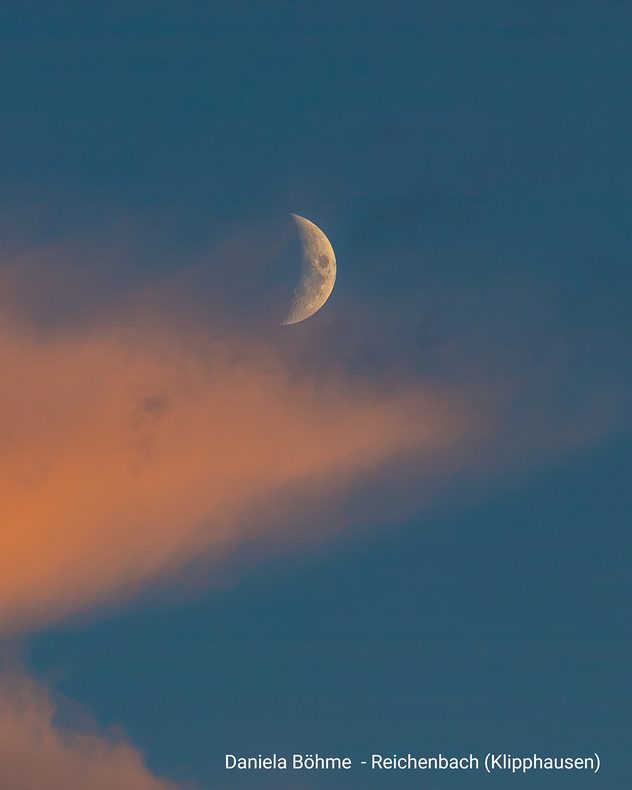 Ein zunehmender Halbmond steht am blauen Abendhimmel über einer rosafarbenen Wolke in der Dämmerung.