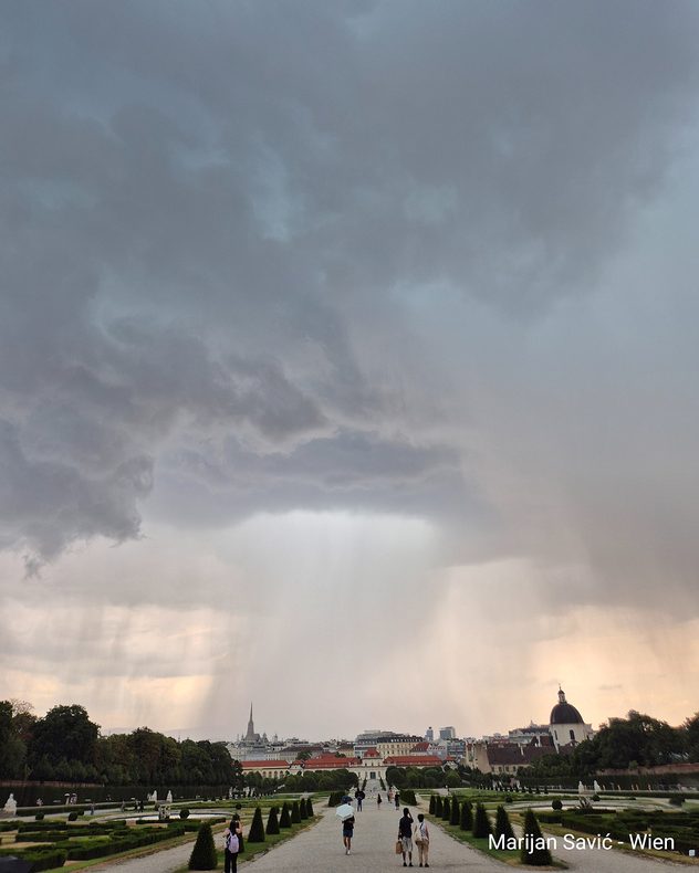 Dunkle Gewitterwolken mit kräftigen Regenfällen ziehen über Wien, während Menschen durch die barocke Parkanlage von Schloss Belvedere gehen.