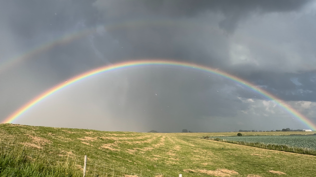 Ein kräftiger doppelter Regenbogen spannt sich unter dunklen Gewitterwolken über eine weite Feldlandschaft nach einem Regenschauer.