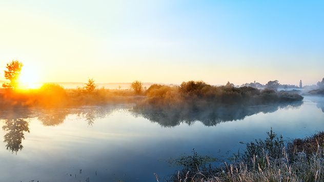 Sonnenaufgang über einem ruhigen Fluss an einem klaren Herbstmorgen. Leichter Nebel steigt über dem Wasser auf, das Ufer ist von Büschen und Gräsern gesäumt – friedliche Hochdruckwetterlage.