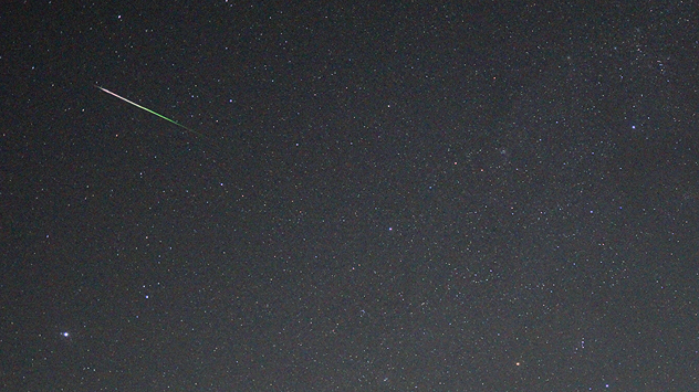 Bright meteor with a white and green trail streaks diagonally across a dark, star-filled night sky.