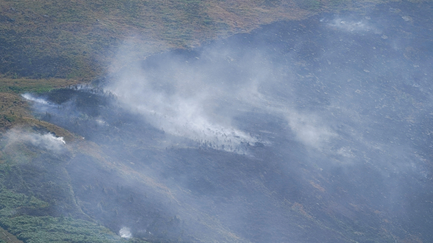 Smoke rising from a scorched moorland hillside after a wildfire, with blackened vegetation, smouldering ground, and drifting gray smoke covering the burned landscape.