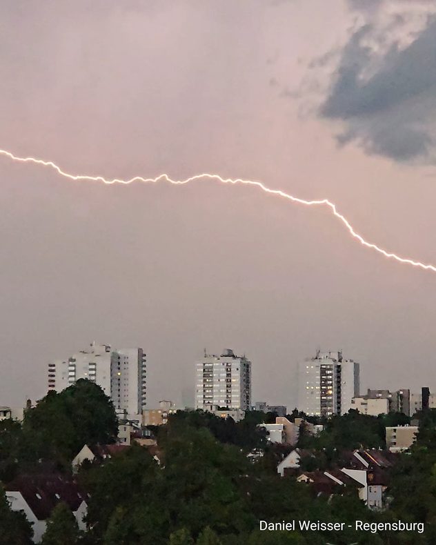 Langer, horizontaler Blitz über einer Stadtsilhouette. Dunkle Wolken und Wohngebäude prägen die Szene.