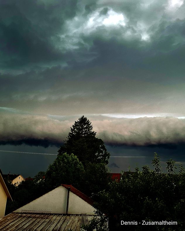 Dunkle Gewitterfront mit markanter Wolkenkante über einer Ortschaft. Im Vordergrund stehen Häuser und hohe Bäume unter bedrohlichem Himmel.