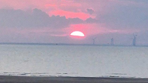 Sunset looking out to the Atlantic Ocean, half hidden behind clouds. A wind farm is seen on the right of the image with the shore running onto the beach at the bottom.