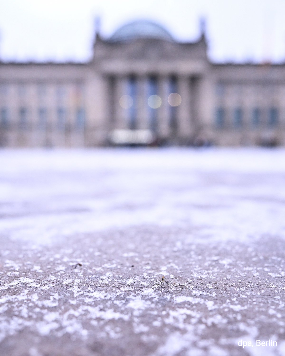 Vereister Boden vor dem Reichstagsgebäude in Berlin, das im Hintergrund unscharf zu sehen ist. Eis- und Schneereste bedecken den Platz.