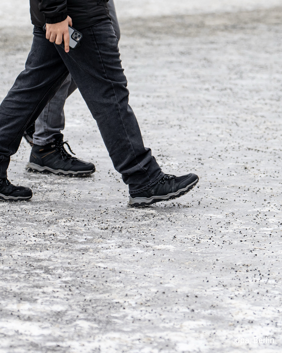 Zwei Fußgänger gehen über eine vereiste Fläche, Streugut liegt auf dem Boden. Winterliche Glätte in der Stadt.