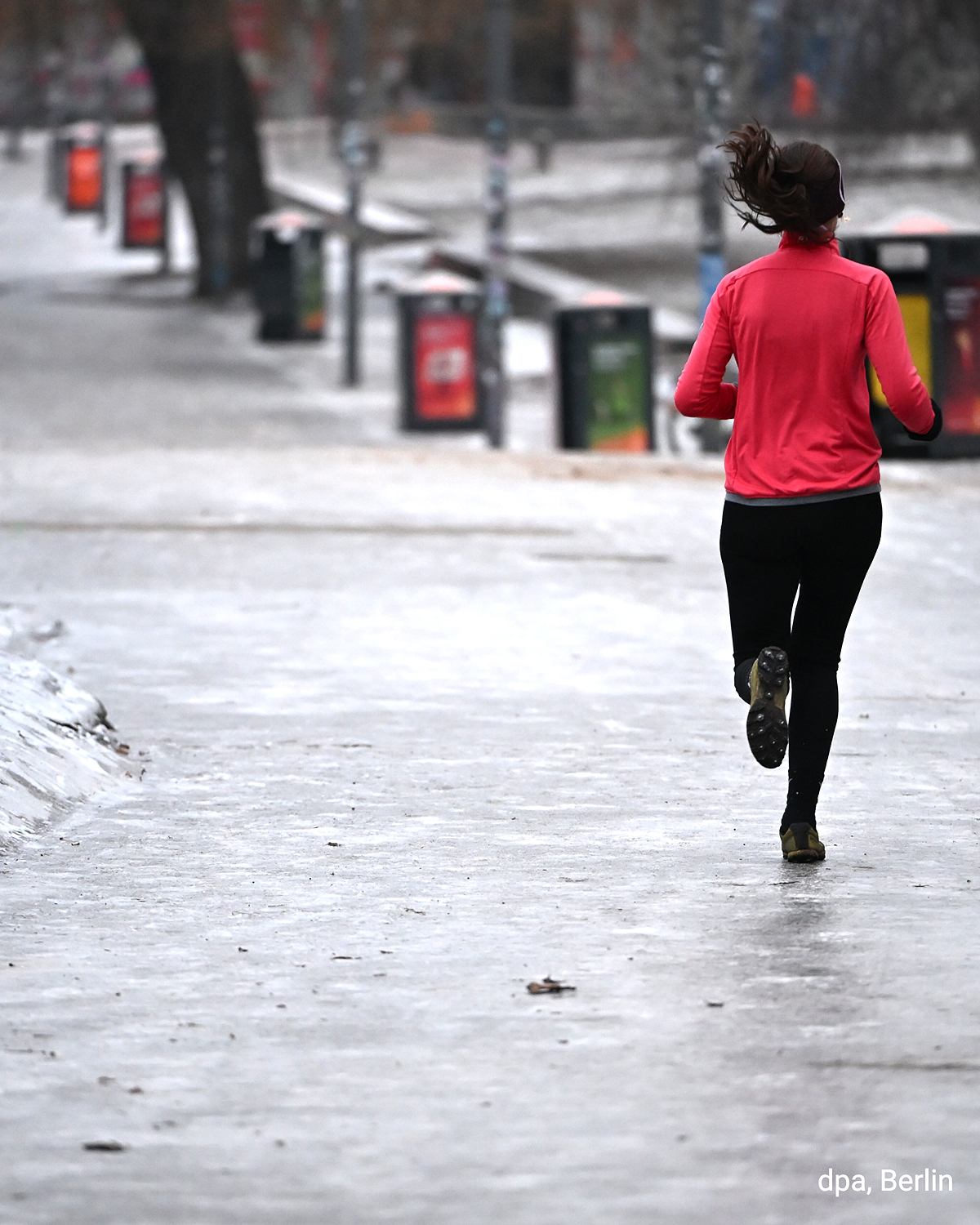 Joggerin in roter Jacke läuft mit Spikes über einen vereisten Weg in Berlin. Glatte Oberfläche und winterliche Umgebung sind erkennbar.