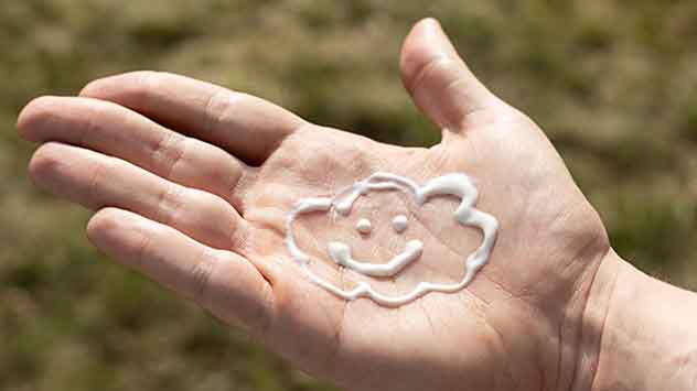 Eine Handfläche mit Sonnencreme, die in Form einer Wolke mit lächelndem Gesicht aufgetragen wurde.