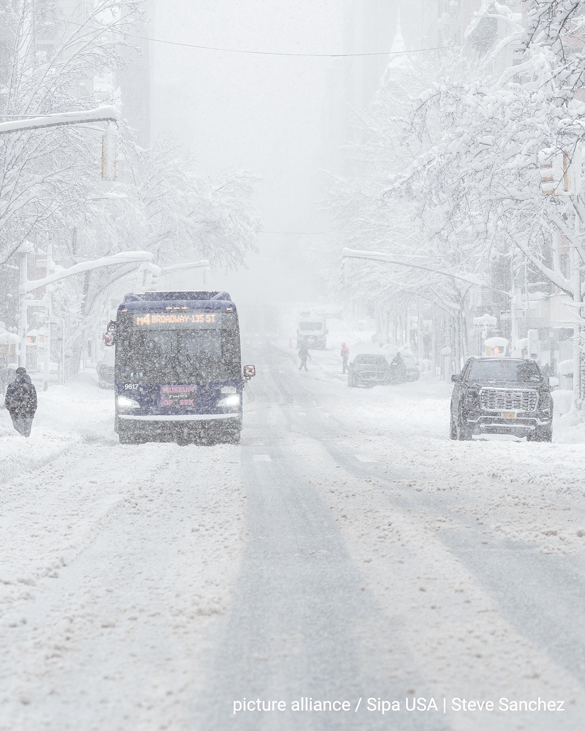 A snow-covered city street on the US East Coast with heavy snowfall. A public bus drives through deep snow, cars are parked at the side of the road. Trees and traffic lights are thickly covered with snow, visibility is severely restricted.