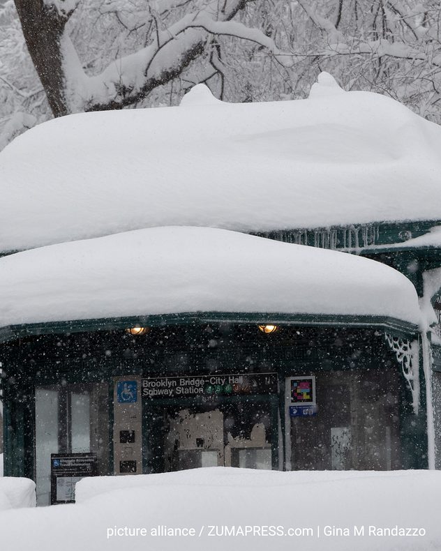 Entrance to the Brooklyn Bridge–City Hall subway station in Manhattan, completely covered in snow. Thick layers of snow lie on the roof and railings, and heavy snowfall reduces visibility. Trees in the background are also heavily iced and snow-covered.