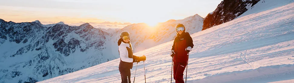 Zwei Skifahrer auf einer Piste bei Sonnenuntergang.