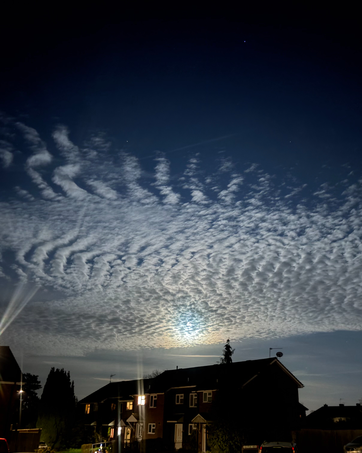 Rippled altocumulus cloud layer lit by moonlight forming a mackerel sky pattern above silhouetted houses, with textured wave-like clouds across a dark blue night sky.