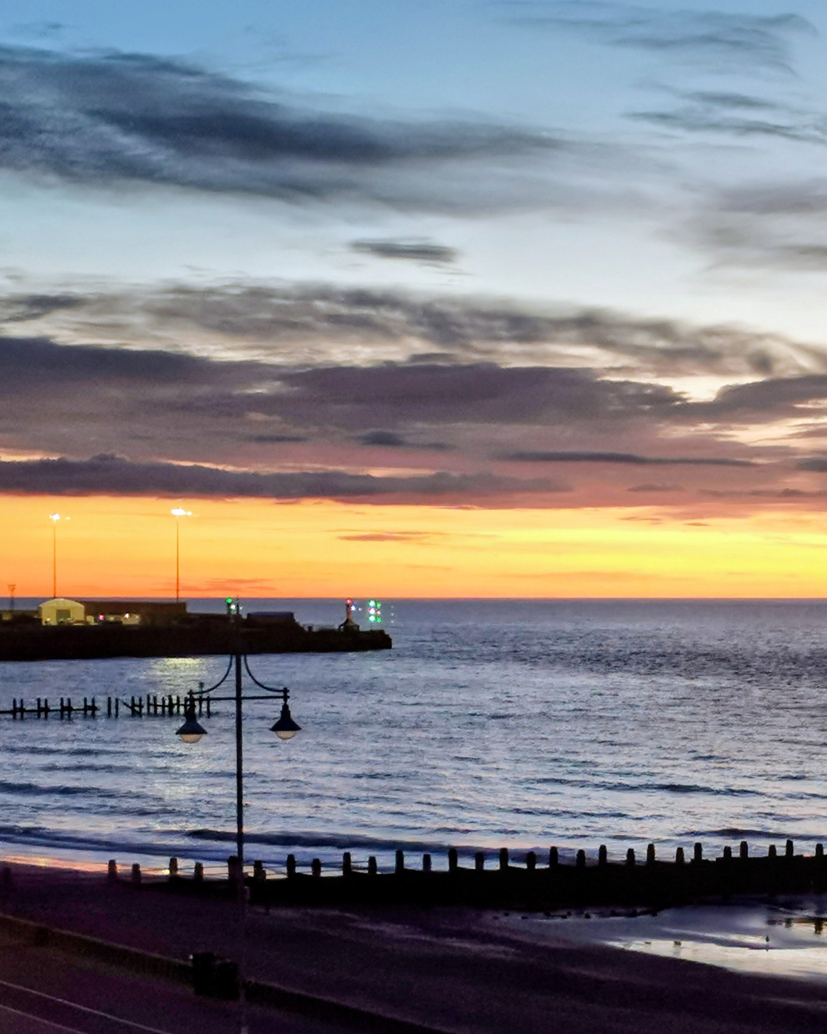 Sunrise over the North Sea at Lowestoft, Suffolk, with an orange and yellow horizon, calm waves, harbour lights in the distance, and silhouetted lampposts and groynes along the shore.