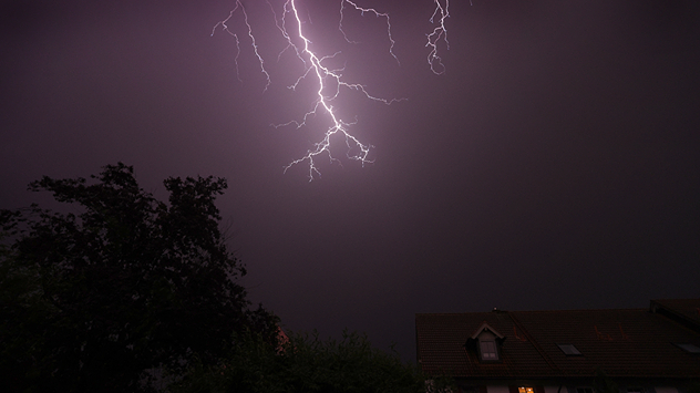 Heller Blitz am Nachthimmel über Häusern und Bäumen. Dunkle Wolken und schwaches Umgebungslicht. Gewitterszene in einer Sommernacht.