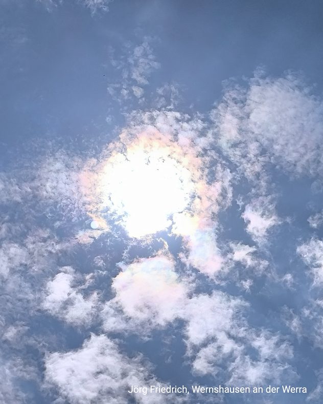Sonne hinter Wolken mit irisierenden Farben in Blau, Rosa und Grün am Himmel.