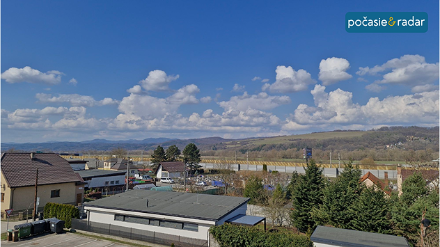 Pohľad na slovenskú krajinu a obec pod modrou oblohou s kopovitými oblakmi.