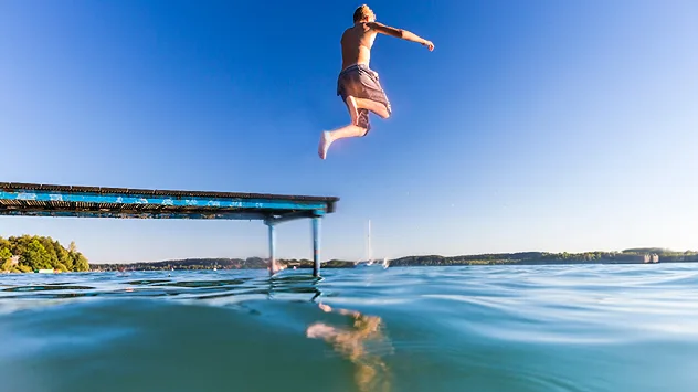 Eine Person springt bei blauem Himmel von einem Steg in einen Badesee. Das Wasser ist ruhig und verspricht an einem sonnigen Sommertag eine erfrischende Abkühlung.