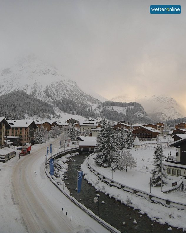 Village alpin enneigé avec montagnes et rivière dans la lumière du matin. Les maisons et les arbres sont profondément enrobés dans la blancheur hivernale.