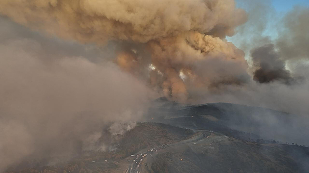 © dpa Luftaufnahme eines großen Waldbrands mit massiven Rauchwolken über hügeligem Gelände und einer Straße mit Fahrzeugen.
